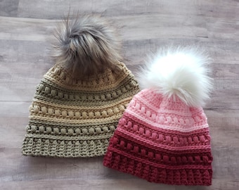 May include: Two handmade crochet beanies with faux fur pom-poms. One beanie is olive green and tan, the other is pink and burgundy. The beanies have a textured stitch pattern and are designed for cold weather.