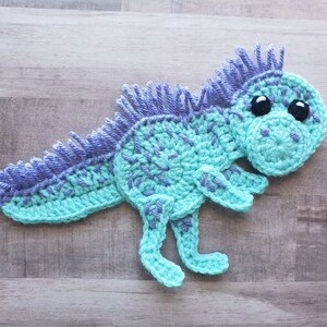 May include: A crocheted dinosaur toy with blue and purple yarn. The dinosaur has large black eyes and a spiky back.