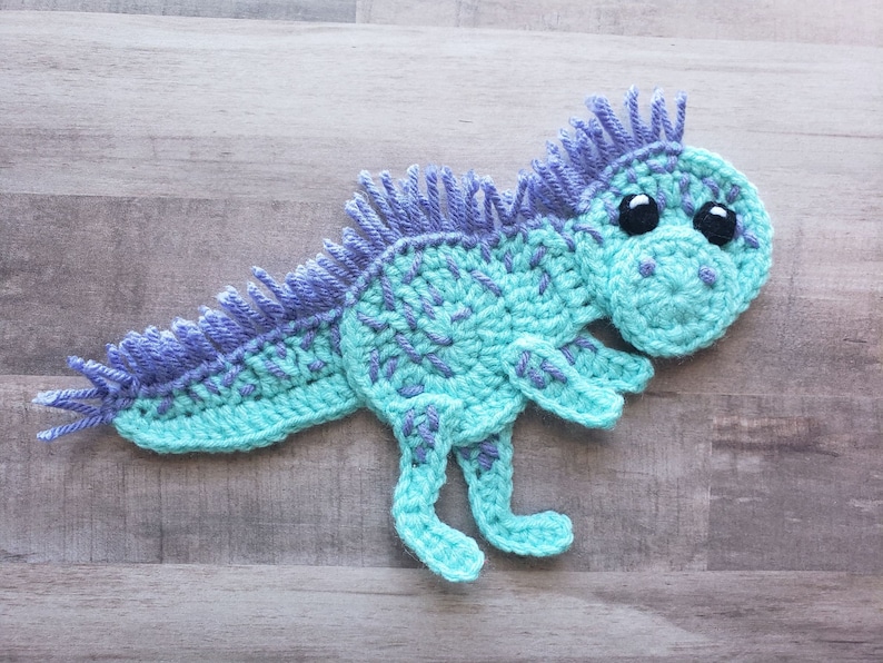 May include: A crocheted dinosaur toy with blue and purple yarn. The dinosaur has large black eyes and a spiky back.