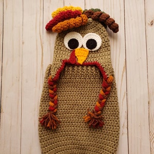 May include: A crocheted baby costume in the shape of a turkey. The costume is brown with a red, orange, and yellow wattle and a brown, orange, and green headpiece. The costume has white eyes and a yellow beak.