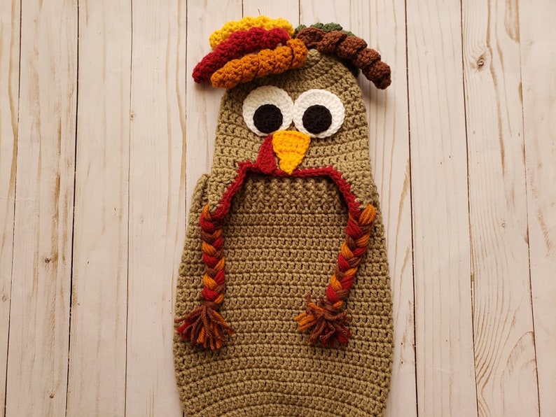 May include: A crocheted baby costume in the shape of a turkey. The costume is brown with a red, orange, and yellow wattle and a brown, orange, and green headpiece. The costume has white eyes and a yellow beak.