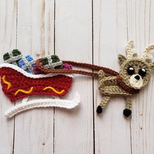 Puede incluir: Un reno de crochet tirando un trineo rojo y blanco lleno de regalos coloridos. El reno tiene pelo marr&oacute;n y blanco, ojos negros y una nariz marr&oacute;n. El trineo tiene una base blanca y un cuerpo rojo con l&iacute;neas onduladas amarillas.