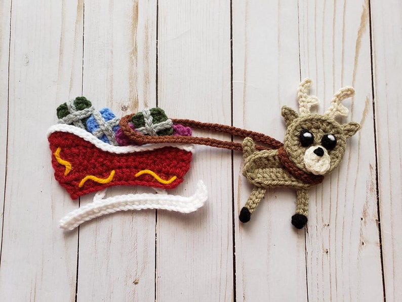 Puede incluir: Un reno de crochet tirando un trineo rojo y blanco lleno de regalos coloridos. El reno tiene pelo marr&oacute;n y blanco, ojos negros y una nariz marr&oacute;n. El trineo tiene una base blanca y un cuerpo rojo con l&iacute;neas onduladas amarillas.