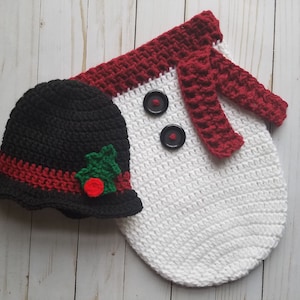 May include: A white crocheted snowman outfit with a red scarf and black buttons. A black crocheted hat with a red band and a green holly accent.