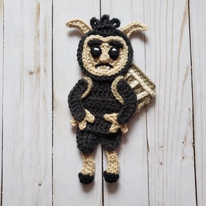 May include: A crocheted amigurumi toy of a creature with horns, a black and brown body, and a white face. The toy is standing on a white wooden surface.