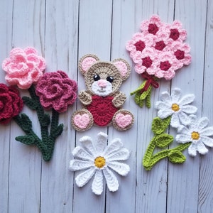 Op de afbeelding: Een set gehaakte bloemen en een teddybeer. De bloemen zijn in tinten roze, rood en wit. De teddybeer is bruin met een rood hart op zijn borst.