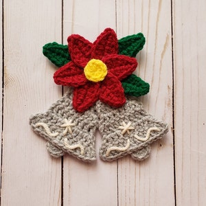 Puede incluir: Dos campanas de ganchillo grises con bordados blancos, coronadas con una flor de poinsettia roja de ganchillo con un centro amarillo y hojas verdes.