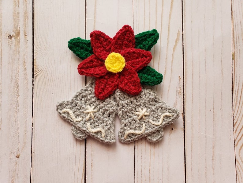 Puede incluir: Dos campanas de ganchillo grises con bordados blancos, coronadas con una flor de poinsettia roja de ganchillo con un centro amarillo y hojas verdes.