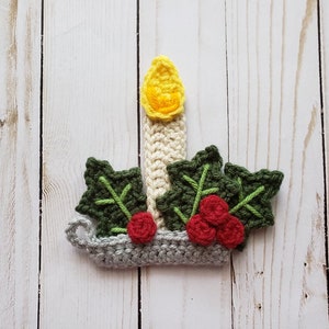 Puede incluir: Un adorno navide&ntilde;o de ganchillo con una vela blanca con una llama amarilla, rodeado de hojas de acebo verdes y bayas rojas.