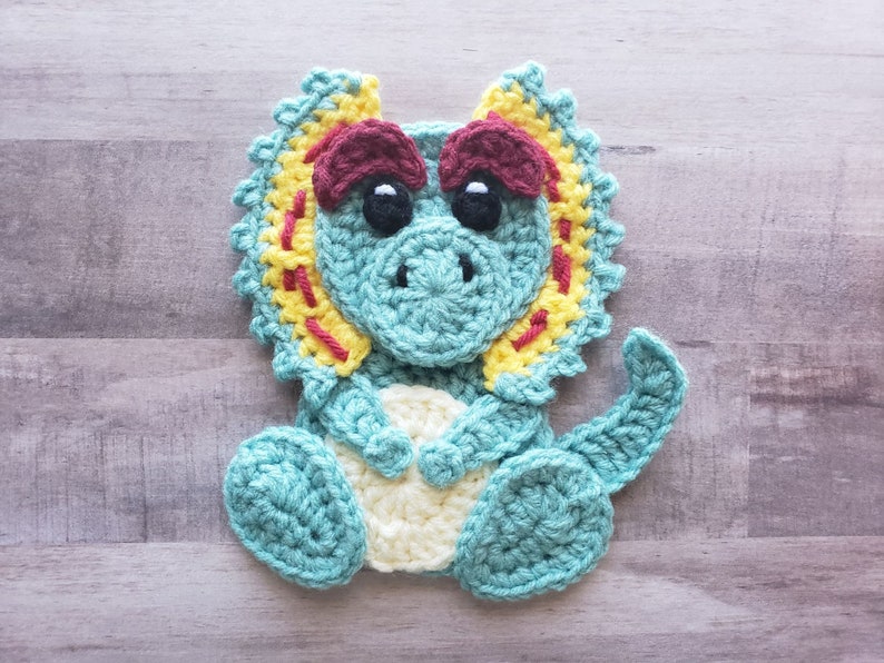 May include: A crocheted triceratops dinosaur toy with a teal body, yellow and red frill, and white eyes.
