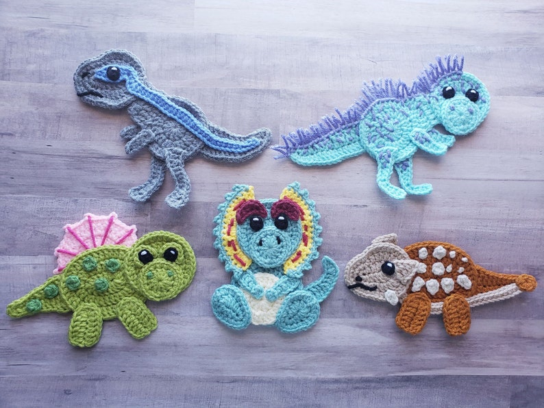 May include: Five crocheted dinosaur toys in different colors and styles. The dinosaurs are a blue and grey T-Rex, a light blue Triceratops, a green Stegosaurus, a light blue Spinosaurus, and a brown Ankylosaurus.