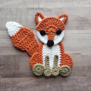 Puede incluir: Una aplicaci&oacute;n de zorro de crochet con hilo naranja, blanco y verde. El zorro tiene una cola blanca, ojos negros y una nariz negra.