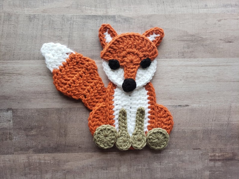 Puede incluir: Una aplicaci&oacute;n de zorro de crochet con hilo naranja, blanco y verde. El zorro tiene una cola blanca, ojos negros y una nariz negra.