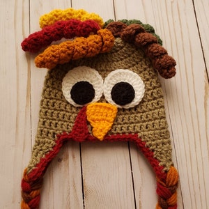 May include: Crocheted turkey hat with brown, orange, red, and yellow yarn. The hat has a brown crocheted body, a red beak, and white eyes with black pupils. The hat has two long brown and orange braids.