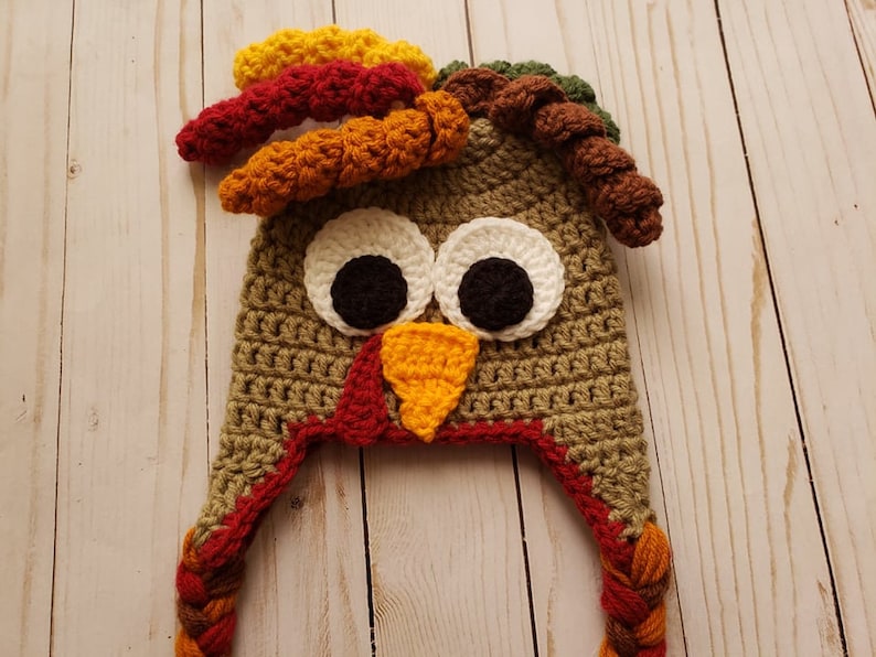 May include: Crocheted turkey hat with brown, orange, red, and yellow yarn. The hat has a brown crocheted body, a red beak, and white eyes with black pupils. The hat has two long brown and orange braids.