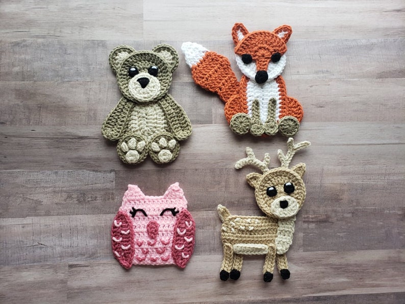Puede incluir: Cuatro aplicaciones de animales de crochet: un oso marr&oacute;n, un zorro rojo, un b&uacute;ho rosa y un ciervo marr&oacute;n. Los animales est&aacute;n todos sentados o de pie sobre un fondo de madera.