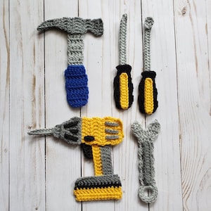 Könnte beinhalten: Eine Sammlung gehäkelter Werkzeuge, darunter ein Hammer mit blauem Griff, zwei Schraubendreher mit schwarzen und gelben Griffen, ein Bohrer und ein Schraubenschlüssel. Die Werkzeuge sind grau, gelb und schwarz.