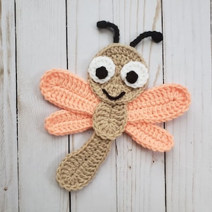 Può includere: Una libellula all'uncinetto con ali di colore marrone e pesca e antenne nere. La libellula ha occhi bianchi e una bocca sorridente.