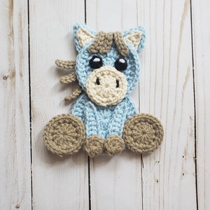 May include: A crocheted blue horse with brown and cream accents. The horse is sitting with its legs crossed and has large black eyes.