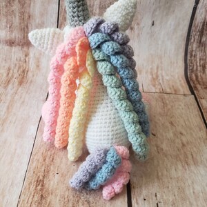 May include: A white crocheted unicorn with a rainbow mane and tail. The unicorn is sitting on a wooden surface.