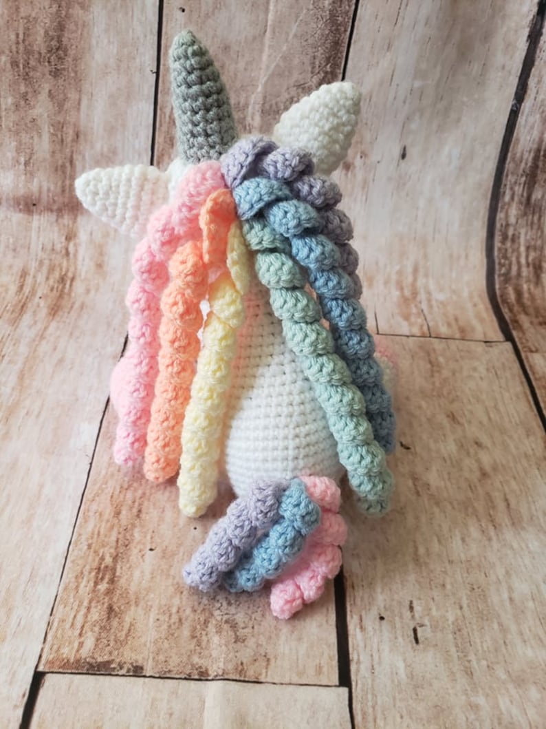 May include: A white crocheted unicorn with a rainbow mane and tail. The unicorn is sitting on a wooden surface.