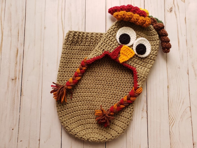 May include: A crocheted turkey costume for a baby, featuring a brown hooded body with a braided tail and a separate turkey head with a red, orange, and yellow wattle, brown eyes, and a yellow beak.