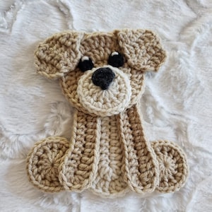Puede incluir: Una aplicación de perro de crochet marrón y beige con ojos negros y una nariz negra.