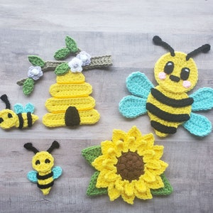Puede incluir: Un conjunto de decoraciones de abeja y girasol de ganchillo. El conjunto incluye una colmena, un girasol y dos abejas. La colmena es amarilla y tiene una entrada marrón. El girasol es amarillo con un centro marrón. Las abejas son amarillas con rayas negras y tienen alas azules.