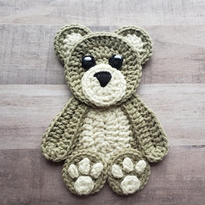 Puede incluir: Una aplicaci&oacute;n de oso de peluche de crochet en tonos verdes y beige con ojos negros. El oso tiene una expresi&oacute;n simple y dulce y es perfecto para agregar un toque de fantas&iacute;a a un proyecto artesanal.