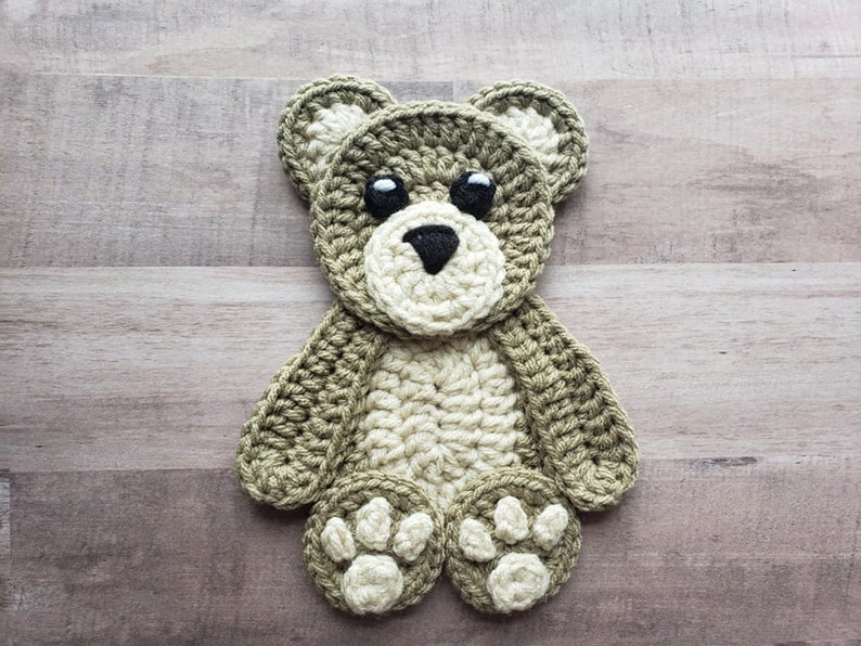 Puede incluir: Una aplicaci&oacute;n de oso de peluche de crochet en tonos verdes y beige con ojos negros. El oso tiene una expresi&oacute;n simple y dulce y es perfecto para agregar un toque de fantas&iacute;a a un proyecto artesanal.