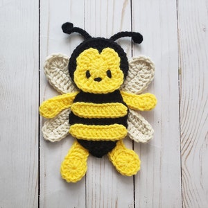 Peut inclure: Une applique de crochet en forme d'abeille avec des rayures noires et jaunes, des ailes blanches et un visage mignon.
