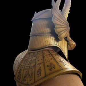 May include: A gold-colored costume helmet with a stylized winged design and a matching shoulder piece with hieroglyphic markings.