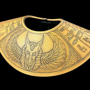 May include: A gold-colored, decorative collar with an Anubis design. The collar is made of a textured material and features intricate details of the Egyptian god Anubis, including his jackal head, wings, and hieroglyphics.