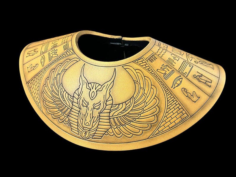 May include: A gold-colored, decorative collar with an Anubis design. The collar is made of a textured material and features intricate details of the Egyptian god Anubis, including his jackal head, wings, and hieroglyphics.