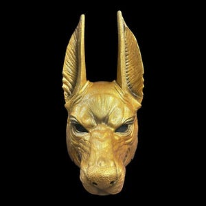 May include: A golden mask of an Anubis jackal head with large ears. The mask is made of a smooth, textured material and has a realistic appearance.