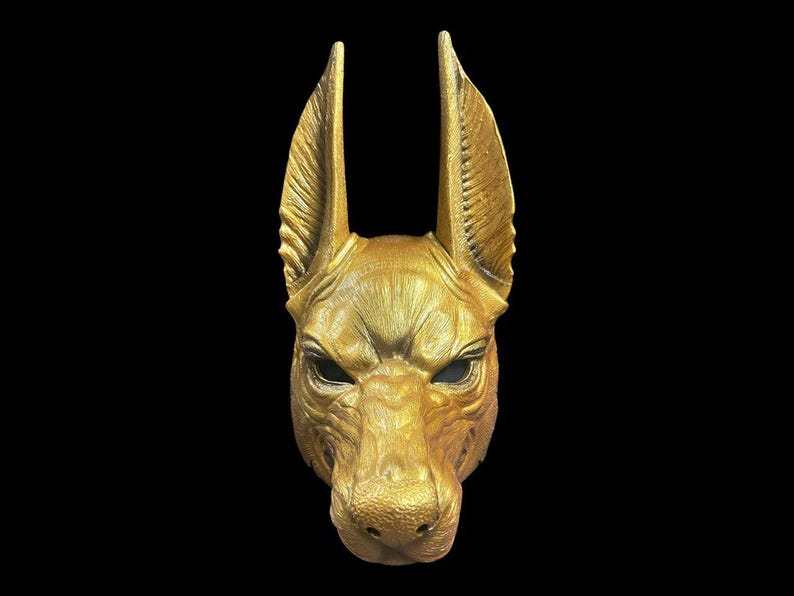 May include: A golden mask of an Anubis jackal head with large ears. The mask is made of a smooth, textured material and has a realistic appearance.