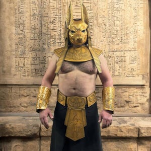 May include: A person wearing a gold Anubis mask, a gold collar, gold arm guards, a gold belt, and a gold kilt. The person is standing in front of a wall with hieroglyphics.