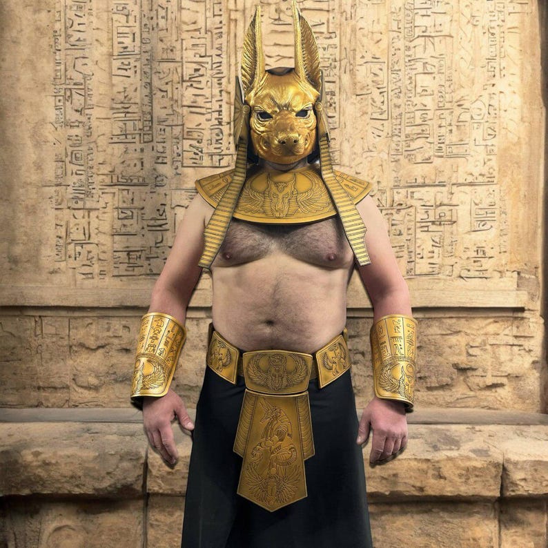 May include: A person wearing a gold Anubis mask, a gold collar, gold arm guards, a gold belt, and a gold kilt. The person is standing in front of a wall with hieroglyphics.