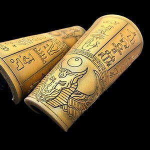 May include: A pair of gold leather arm cuffs with an Egyptian-inspired design. The cuffs feature hieroglyphics and a winged ankh symbol.