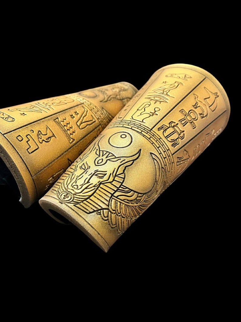 May include: A pair of gold leather arm cuffs with an Egyptian-inspired design. The cuffs feature hieroglyphics and a winged ankh symbol.
