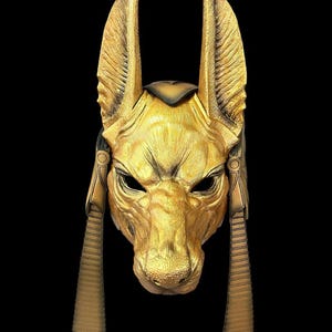May include: A gold-colored mask with a jackal head design. The mask has large ears, a pointed snout, and a detailed face. The mask is decorated with gold accents and has a textured surface.