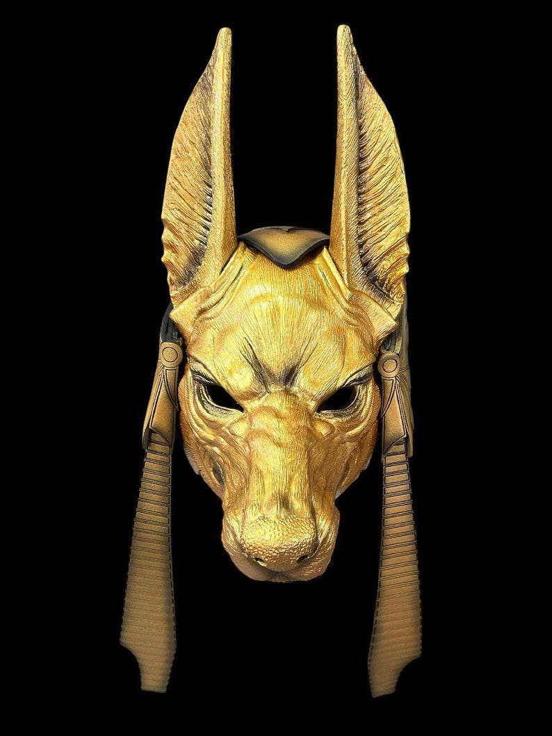 May include: A gold-colored mask with a jackal head design. The mask has large ears, a pointed snout, and a detailed face. The mask is decorated with gold accents and has a textured surface.