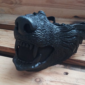 Può includere: Una maschera da lupo nera con un design dettagliato e testurizzato. La maschera ha una bocca spalancata con denti affilati e una rete nera che copre l'interno della bocca.