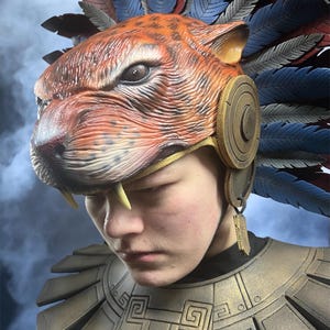 May include: A detailed close-up of a jaguar-themed headdress with a feathered crown. The headdress is predominantly orange with black spots, gold accents, and blue and red feathers. The person is wearing a gold-colored shoulder armor.