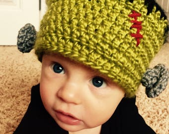 May include: A lime green and black crocheted beanie hat designed to resemble a monster. The hat features a black top section, two grey crocheted bolts attached to the sides, and a red stitched scar detail on the front left side of the hat.