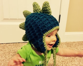 May include: A baby wearing a green and blue crocheted dinosaur hat with long green and blue tassels. The hat has a textured design and a pointed top.