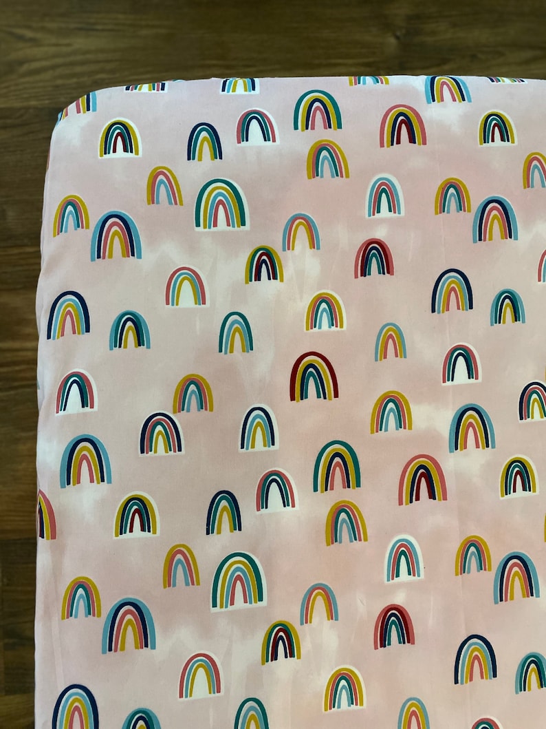 May include: Pink fitted sheet with a repeating pattern of colorful rainbows. The sheet is made of soft, breathable fabric and is perfect for a crib or toddler bed.