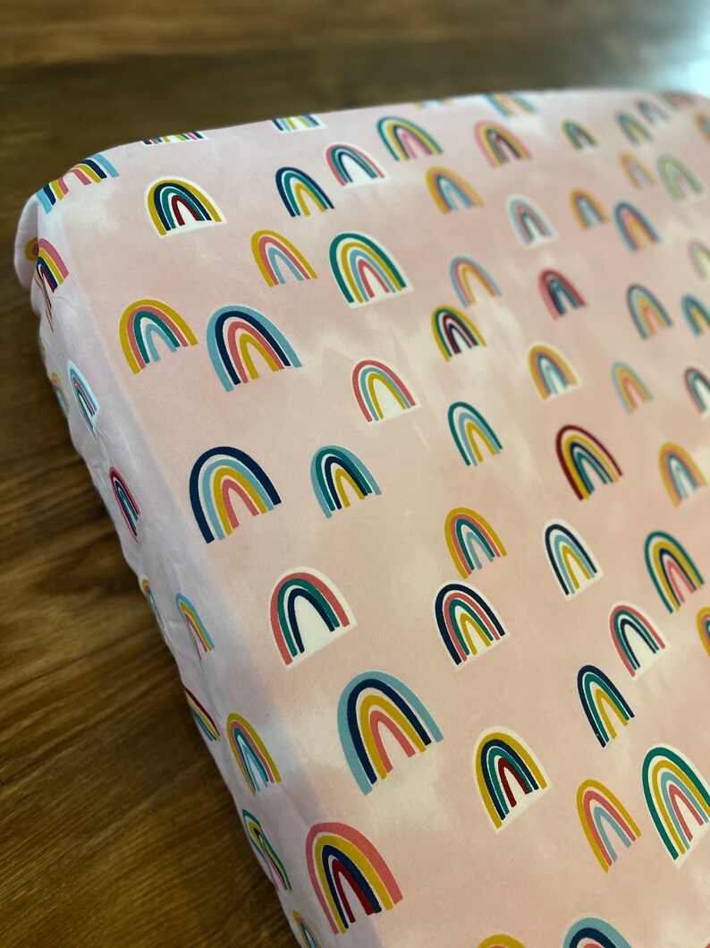 May include: Pink fitted crib sheet with a repeating pattern of colorful rainbows.