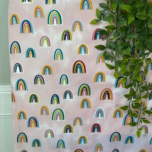 May include: Pink fabric with a repeating pattern of colorful rainbows. The rainbows are in various shades of blue, green, yellow, red, and white.
