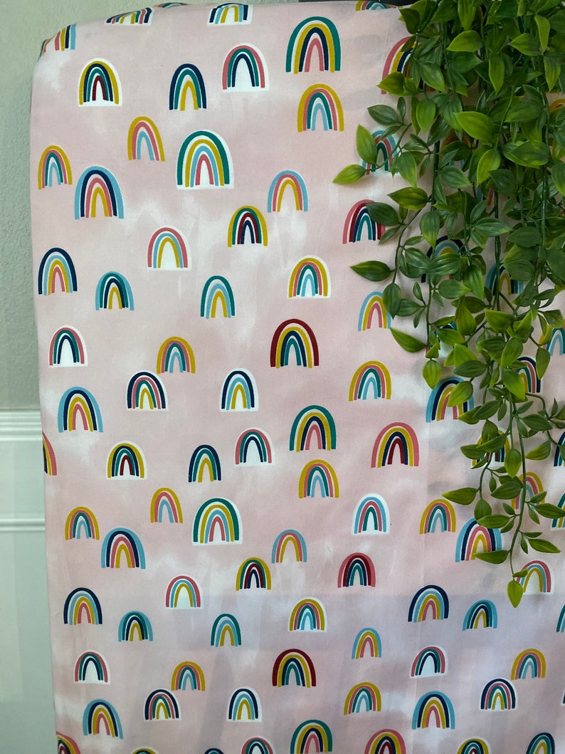 May include: Pink fabric with a repeating pattern of colorful rainbows. The rainbows are in various shades of blue, green, yellow, red, and white.